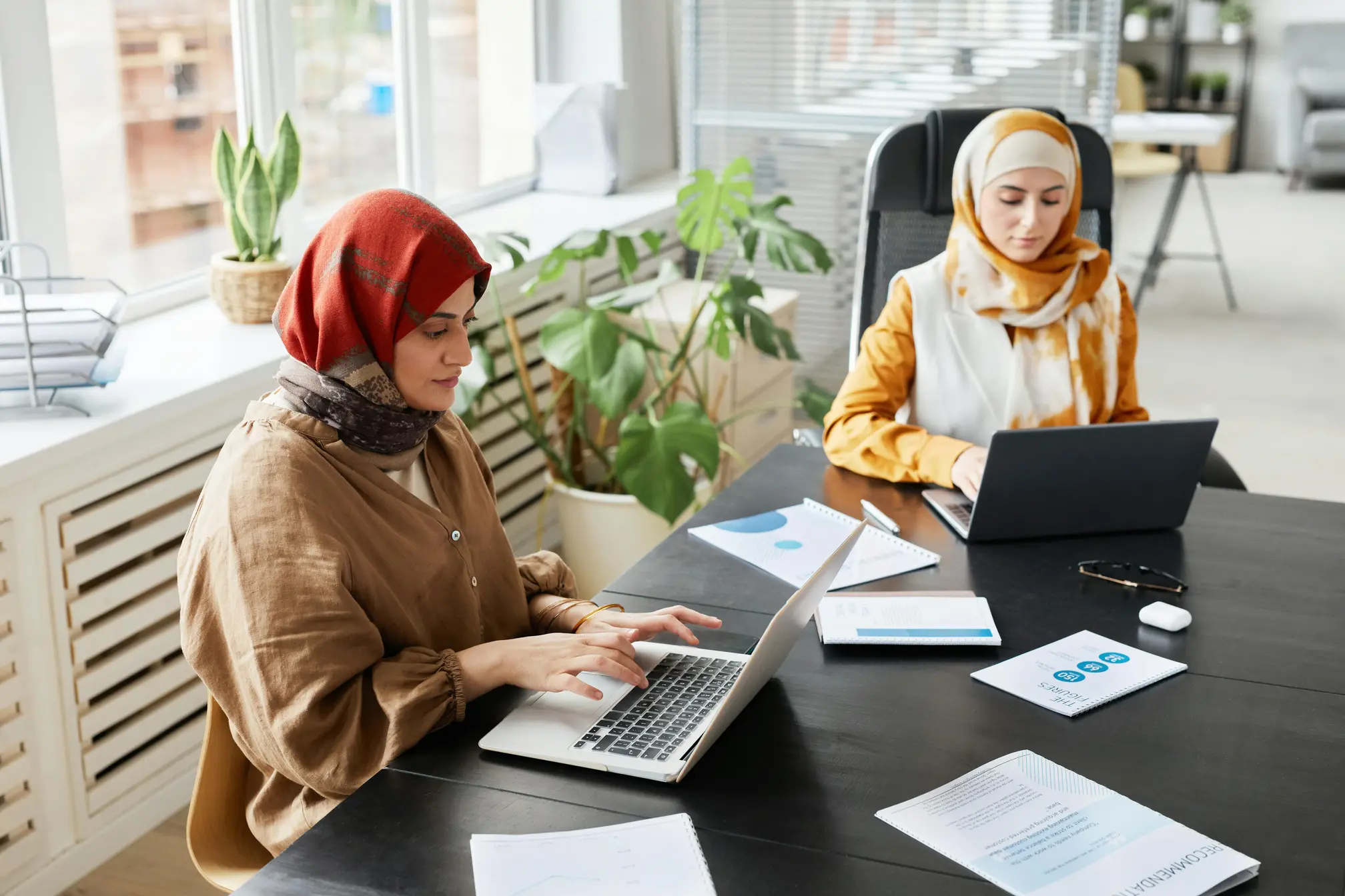 Two Muslim women wearing hijabs sit around a conference room table working in translator jobs on their laptops at Aurora Hikma.